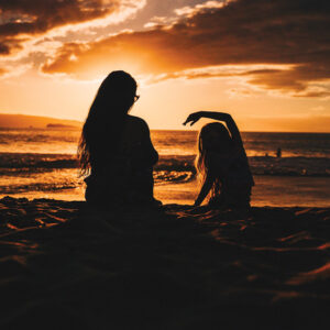 A woman and a girl sitting on the beach together at sunset